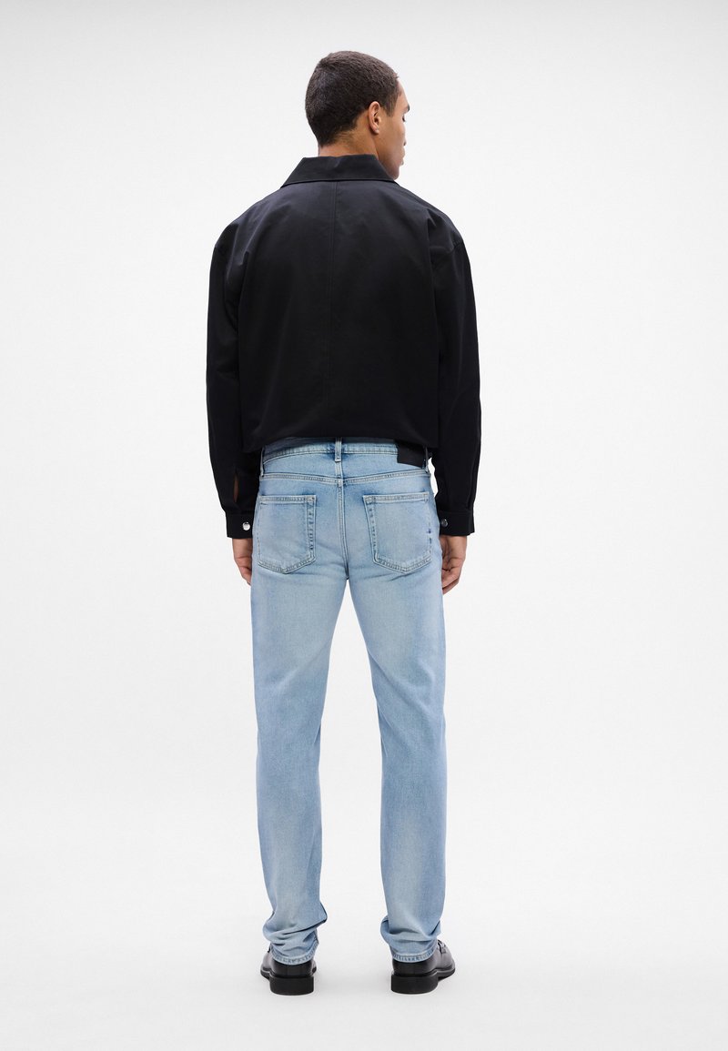 Black, long-sleeve shirt with a collar, paired with light blue jeans and black shoes, viewed from the back.