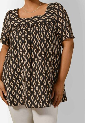 Frau trägt eine kurzärmelige Bluse mit schwarz-beigem Muster und quadratischem Ausschnitt sowie hellfarbene Hose vor neutralem Hintergrund.