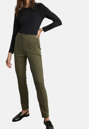 Frau trägt schwarzen Langarm-Top, olivgrüne, hoch taillierte Hose mit Frontnähten und schwarze Leder-Loafers vor einfarbigem Hintergrund.