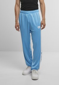 Hellblaue Jogginghosen mit elastischem Bund, weißen Seitenpaneelen und einem Logodetail am linken Oberschenkel. Kombiniert mit weißen Sneakers.