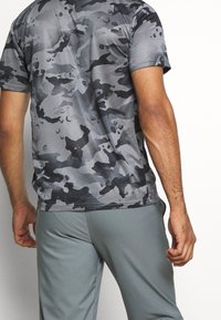 Grå, camouflagemønstret kortærmet skjorte i let stof, kombineret med grå atletiske shorts. Subtil tekstur og moderne pasform er synlig.