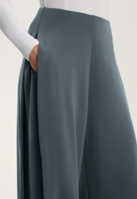 Široke hlače v zatemnjeni teal barvi, s proširitvami, gladko teksturo in stranskimi žepi. Pas je visok in oprijet.