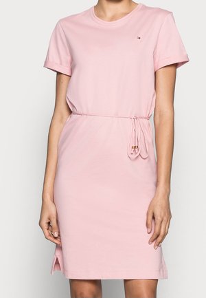 Kobieta ubrana w różową sukienkę do kolan z krótkim rękawem, z wiązaniem w pasie i małym rozcięciem po boku, ozdobioną małym logo na piersi.