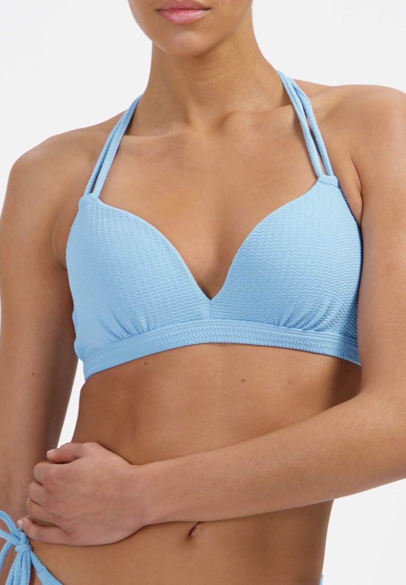 Žena oblečená v texturovaném světle modrém halter bikinovém topu s dvojitými ramínky, ruka opřená o pas na bílém pozadí.