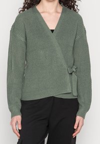 Kvinna i en strukturert olivgrön omslagskofta knuten i sidan, tillsammans med svarta byxor mot en neutral bakgrund.