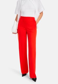 Rote weite Hose aus glattem Stoff, mit hoher Taille und Seitentaschen, kombiniert mit einem weißen strukturierten Oberteil und schwarzen Schuhen.