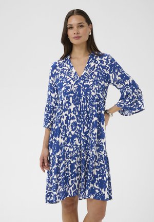 Kobieta o ciemnych włosach, ubrana w niebiesko-białą kwiecistą sukienkę do kolan z rękawami do łokci, stojąca z jedną ręką na biodrze.