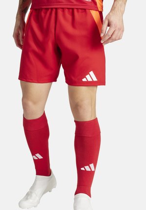 Rode sportshorts met verticale ribbels, wit logo, gecombineerd met rode kniekousen met witte accenten en witte voetbalschoenen.