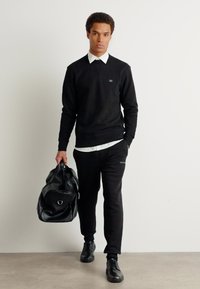 Sweater preto de mangas longas por cima de uma camisa branca com colarinho, combinado com calças de moletom pretas e sapatilhas pretas. A segurar uma grande mala de viagem preta.