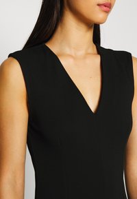 Kvinna i en ärmlös svart klänning med en djup V-ringning som visar halslinjen, axlarna och en del av den övre bröstkorgen.