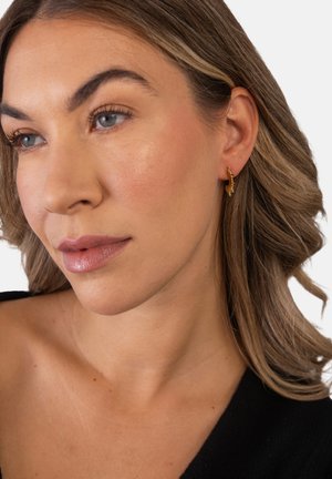 Woman with light brown wavy hair, wearing small gold hoop earrings and a black off-shoulder top, looking slightly left with neutral expression.