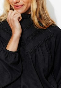 Blouse noire avec un col haut à volants, dotée d'un tissu texturé et de délicats détails brodés sur la poitrine et les manches.