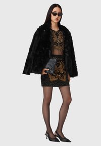 Schwarzer Kunstpelzmantel, durchsichtiges Oberteil mit goldenen Verzierungen, schwarzer Perlenrock, Clutch, schwarze Sonnenbrille und schwarze High Heels.