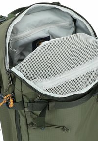 Interior de una mochila de tela verde con un forro gris, un bolsillo de malla y un compartimento con cremallera. Incluye correas ajustables y accesorios.
