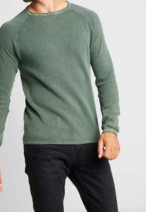 Homme portant un t-shirt vert texturé à manches longues et col rond, ainsi qu'un pantalon noir, debout devant un fond uni gris clair.