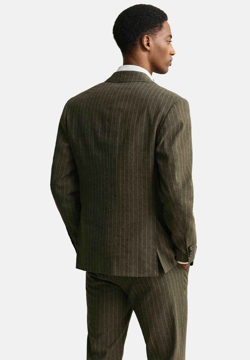 Man staat met handen in de zakken, draagt een donkerbruin krijtstreep pak en een wit overhemd, kijkt naar rechts tegen een effen achtergrond.