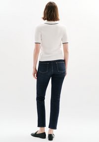 Witte polo met korte mouwen en zwarte afwerking, donkere slim-fit jeans en zwarte instappers, gezien vanuit de achterkant tegen een lichte achtergrond.