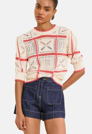 Kobieta nosząca biały sweter z dzianiny z koralowym wzorem w kratkę i bufiastymi rękawami, zestawiony z ciemnymi dżinsowymi szortami z przednimi kieszeniami.