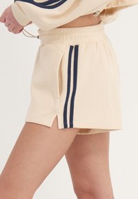 Pantalones cortos de punto waffle beige con franjas laterales en azul marino, equipados con cintura de cordón y bolsillos laterales, diseñados con un dobladillo ligeramente curvado.