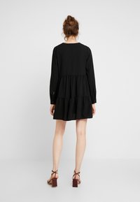 Schwarzes langärmeliges Kleid mit einem gestuften Rock, aus leichtem Stoff. Verfügt über einen runden Halsausschnitt und Knopfmanschetten.