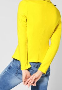 Světlá žlutá, úzká bunda s dlouhými rukávy a vysokým límcem. Švy se táhnou dolů po zádech. V kombinaci s modrými džínami.