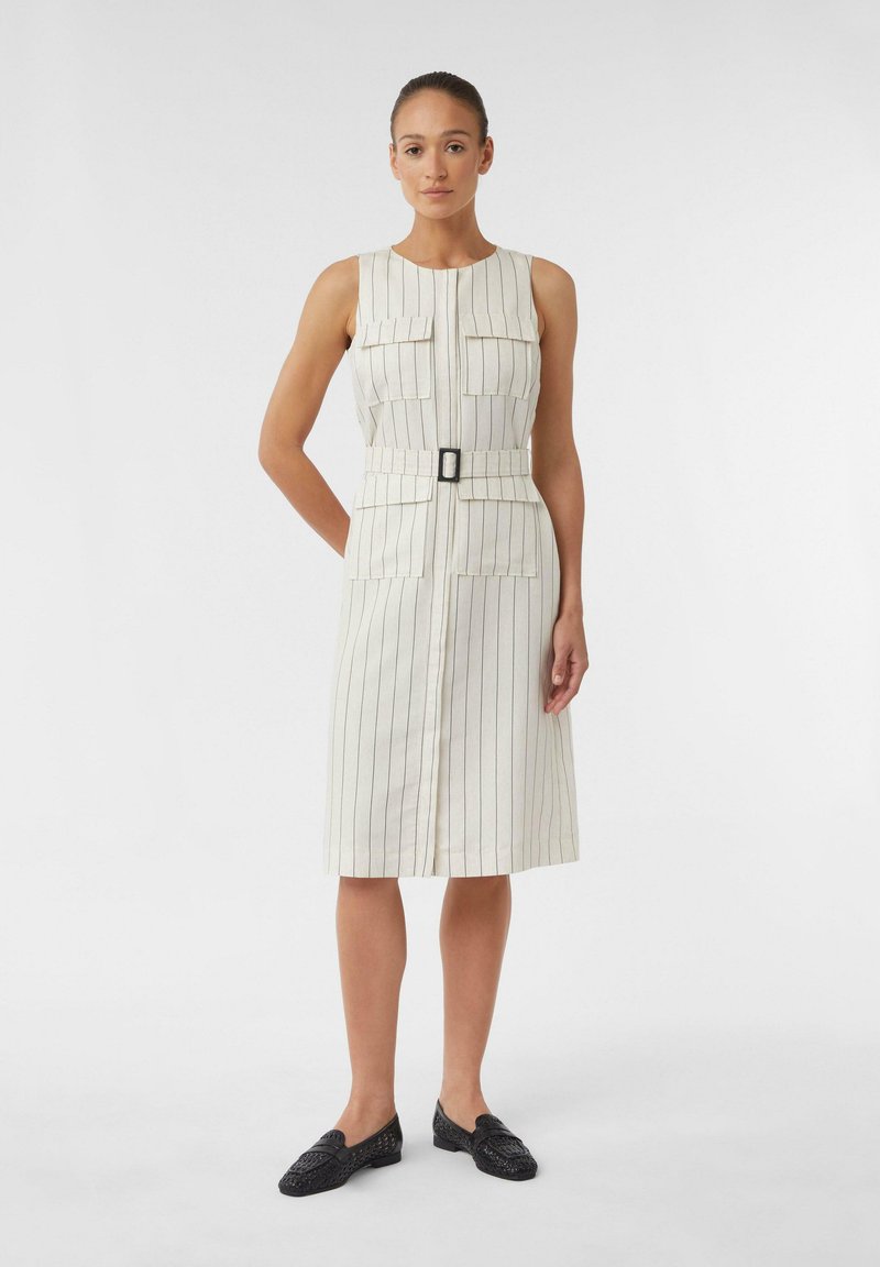 Beżowa i biała sukienka bez rękawów w paski z dwiema przednimi kieszeniami, przewiązana w talii i o prostym kroju. Zestawiona z czarnymi butami wsuwanymi.