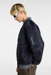 Die marineblaue Bomberjacke verfügt über eine mit Reißverschluss verschlossene Ärmeltasche, gerippte Bündchen und einen leichten Stoff. Das Modell trägt graue Hose und weiße Sonnenbrille.