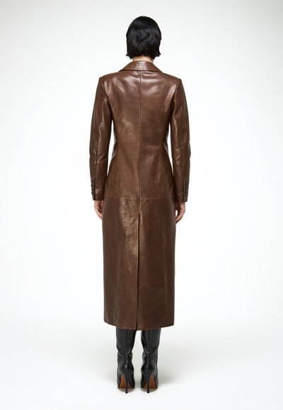 Személy hosszú barna bőrkabátot és fekete sarkú csizmát viselve, háttal állva fehér háttéren.