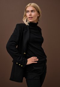 Šviesiaplaukė moteris juodu golfu, švarku su auksiniais mygtukais ir kelnėmis, pozavusi ranką laikydama ant klubo prieš rudą foną.