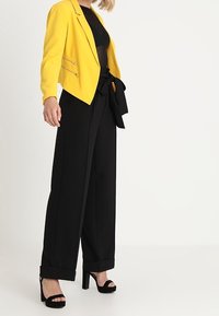 Žlutý blazer se zipovými detaily, černý top, černé kalhoty s vysokým pasem a širokými nohavicemi a černé sandály na vysokém podpatku. Hladká tkanina, módní střih.
