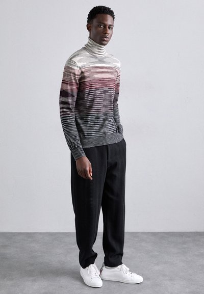 Juostuotas kaklo megztinis pilkų, rožinių ir baltų atspalvių, derinamas su juodomis zigzaginiomis kelnėmis ir baltomis sportinėmis bateliais.