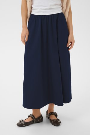 Femme portant une longue jupe bleu marine et des ballerines Mary Jane noires avec des boucles argentées, debout devant un fond blanc uni.