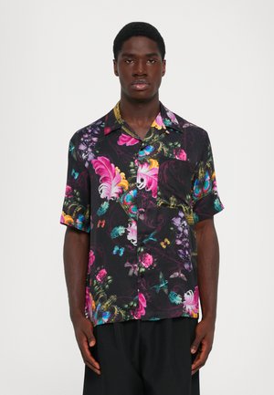 Dark short-sleeve button-up shirt with colorful floral, feather, butterfly, and bird patterns worn by a standing person against a plain background.