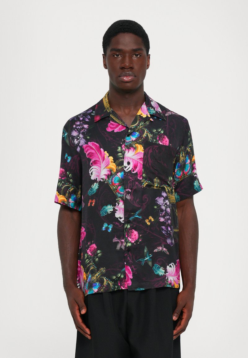 Chemise sombre à manches courtes avec des motifs floraux, plumes, papillons et oiseaux colorés portée par une personne debout devant un fond uni.