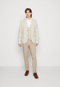 Ljust beige linnekostym med en figuret blazer, väst och byxor, kompletterad med en vit skjorta och bruna läderskor.
