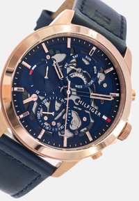 Montre en or rose avec un cadran squelette bleu, comportant des marqueurs de jour, trois sous-cadrans et un bracelet en cuir bleu marine. Fins accents métalliques présents.