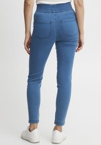 Pantalones de denim azules con una cinturilla elástica alta, dos bolsillos traseros y un corte ajustado. Combinados con zapatillas blancas, mostrando los dobladillos doblados.