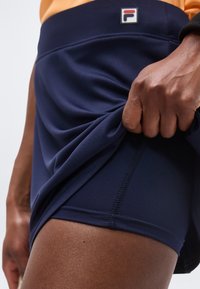 Κοντινό πλάνο ενός ατόμου που σηκώνει το τελείωμα των ναυτικών μπλε αθλητικών σορτς, αποκαλύπτοντας στενά εφαρμοστά εσωτερικά συμπιεστικά σορτς και μια μέση με λογότυπο.