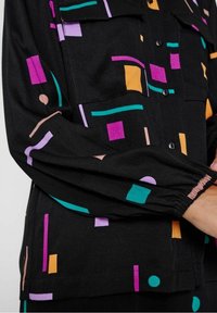 Černá košile s barevnými geometrickými vzory v purpurové, tyrkysové, oranžové a růžové. Obsahuje zapínání na knoflíky a elastické manžety na rukávech.