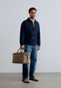 Άνδρας που φοράει μπλε σκούρο σακάκι, λευκό πουκάμισο, μπλε τζιν και καφέ παπούτσια, κρατώντας μια μεγάλη καφέ δερμάτινη τσάντα δίπλα του.