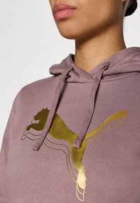 Violeta kapuce ar lielu zelta Puma logo. Dizainam ir teksturēta apdare ar auklām ap kaklu.