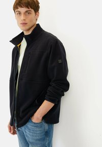 Schwarze Fleecejacke mit hohem Kragen, Frontreißverschluss, linke Brusttasche und seitlichen Taschen. Getragen über einem hellgrünen Hemd und kombiniert mit blauen Jeans.