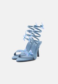 Μπλε ψηλοτάκουνα σανδάλια με σπειροειδείς λουριά στον αστράγαλο, στολισμένα με διαφανείς μπλε πεταλουδένιες διακοσμήσεις σε απλό φωτεινό φόντο.