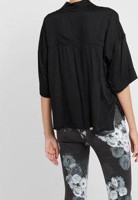 Zwarte, loszittende blouse met een kraag en korte mouwen. Glad materiaal, boxy ontwerp. Gecombineerd met donkere, bloemenpatroon legging.