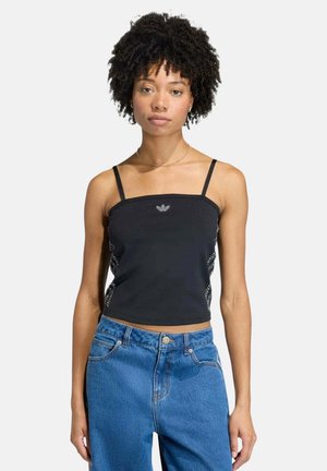 Giovane donna con capelli ricci che indossa un top nero con spalline sottili e jeans blu a vita alta, in piedi davanti a uno sfondo semplice.