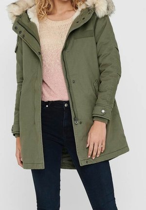 Femme portant un parka vert olive avec capuche en fourrure, un pull en maille rose clair et un jean bleu foncé, debout avec la main gauche le long du corps.