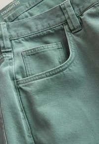 Gros plan de jeans en denim vert montrant la poche avant, les passants de ceinture et les coutures avec une étiquette portant le texte "STRAIGHT FIT".