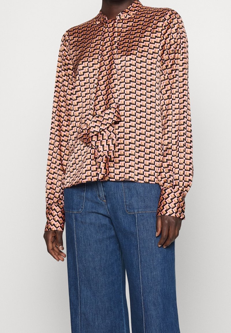 Personne portant un chemisier à motifs géométriques rose et noir avec des volants et un jean bleu taille haute à jambes larges sur un fond uni.
