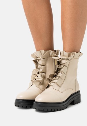 Bottines beiges en cuir, avec des bords volantés, un laçage sur le devant, et une semelle noire épaisse pour le contraste.