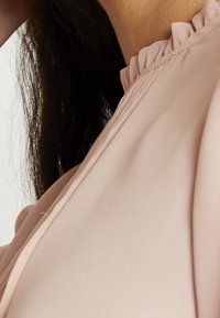 Nahaufnahme des Nackens und der Schulter einer Person mit langen dunklen Haaren, die eine blassrosa Bluse mit einem Rüschensaum trägt.